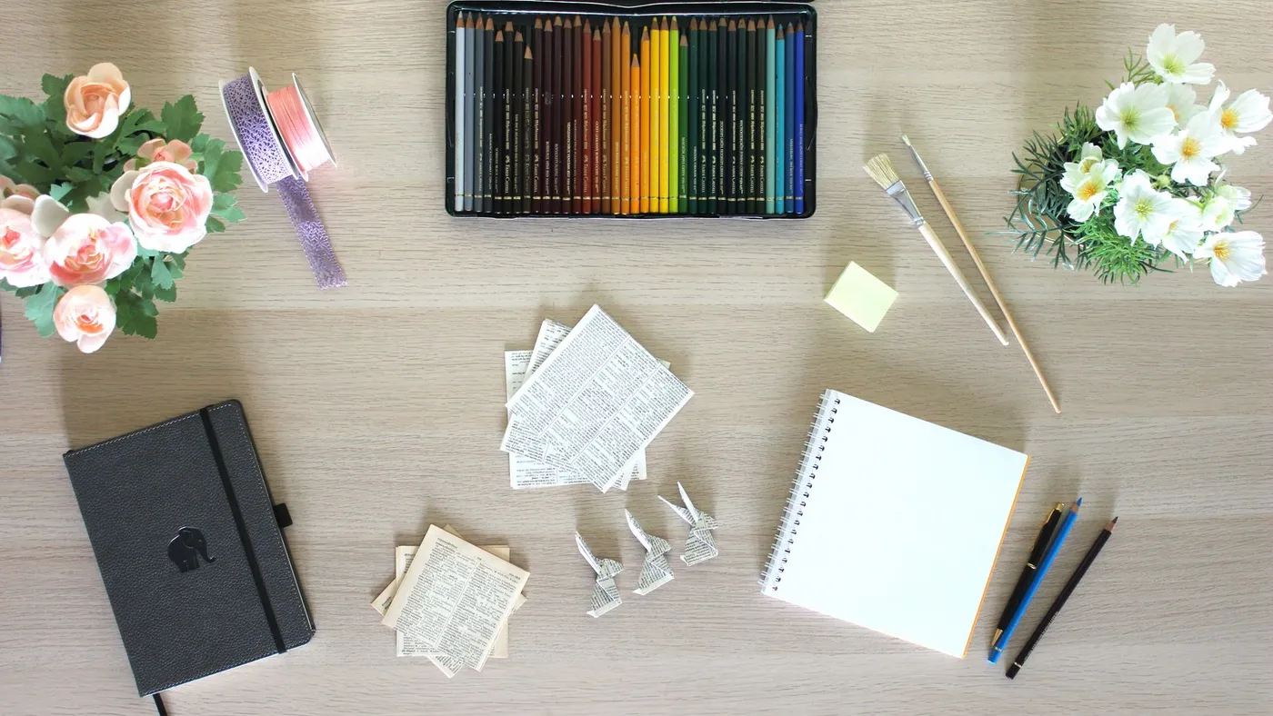 Colored pencil set, notebooks, flowers, and small paper-folded figures on a wooden desk